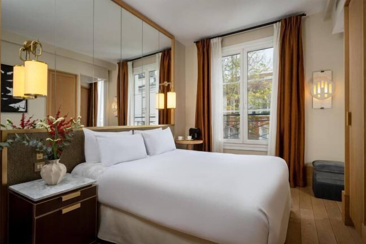 A hotel room with a large bed, a bedside table with a vase, a mirror, a window with curtains, and a balcony view.