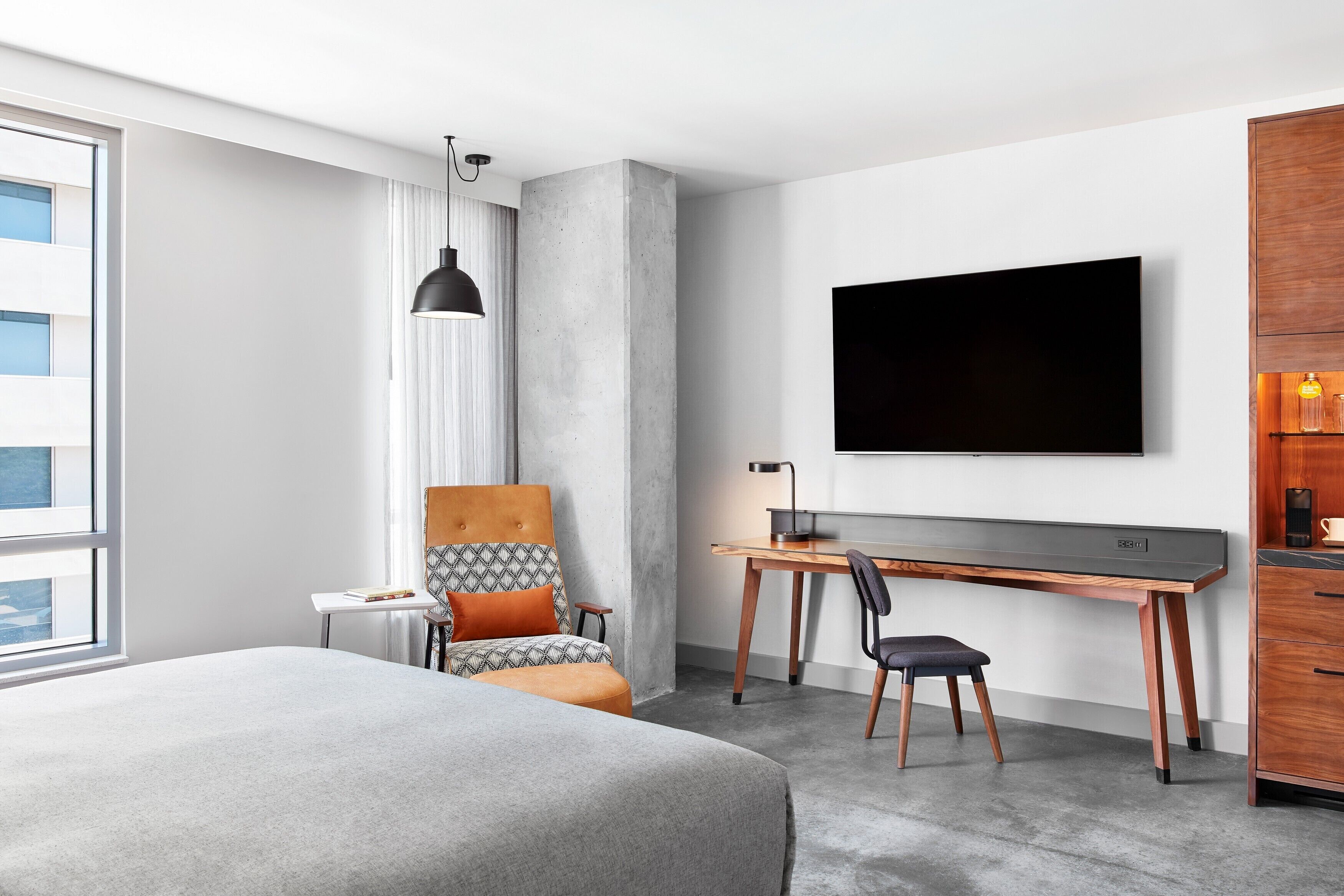 A modern hotel room with a large bed, a desk with a computer, a TV mounted on the wall, and a large window.