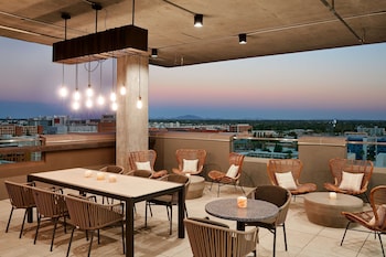 A modern bar area with a high ceiling, a central bar counter, and a seating area with wicker chairs and a sofa. at Canopy by Hilton Tempe Downtown