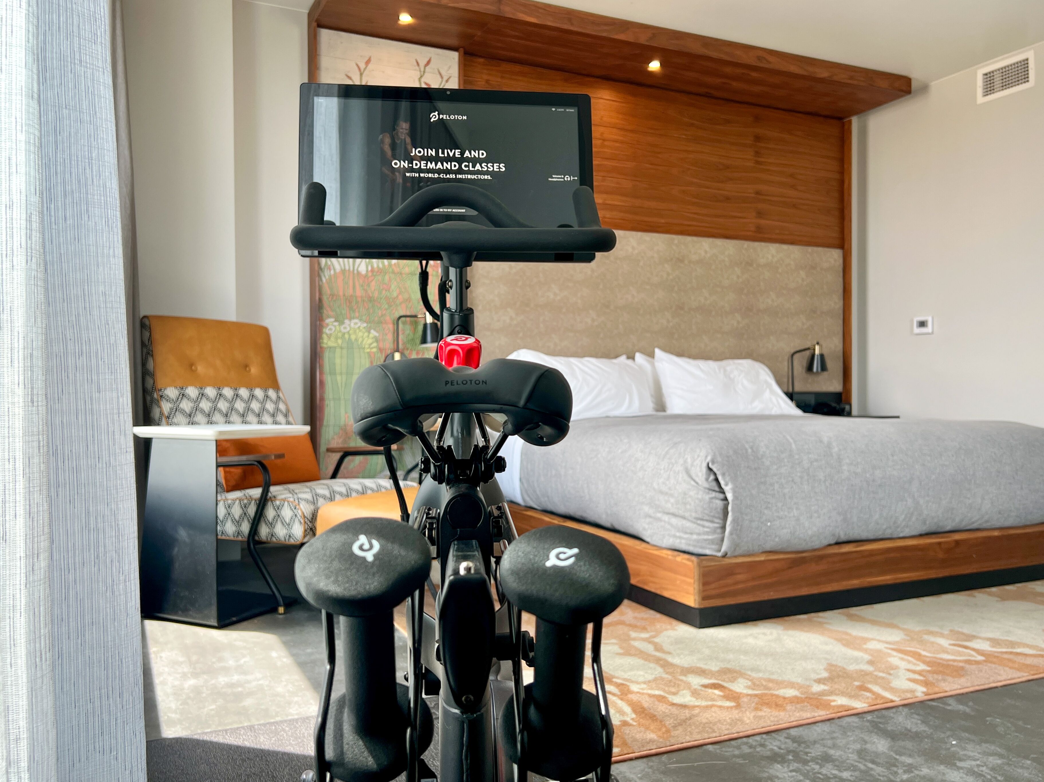 A Peloton bike in a hotel room with a bed, a desk, and a TV displaying Peloton's live and on-demand classes.