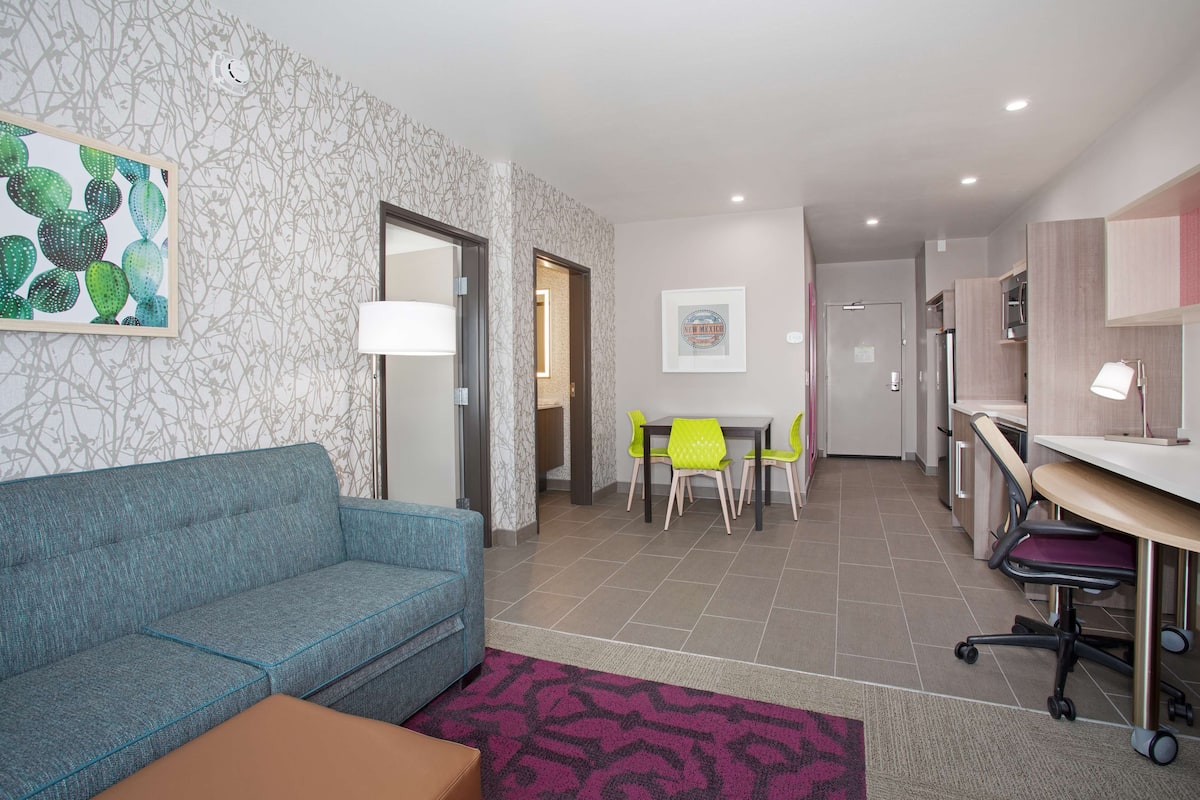 A modern hotel room with a blue sofa, a dining table with yellow chairs, a desk with a chair, and a patterned rug.