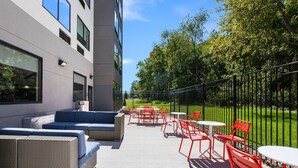 Een terras buiten met zitplaatsen en tafels, een modern gebouw met grote ramen, en een groen gazon.
