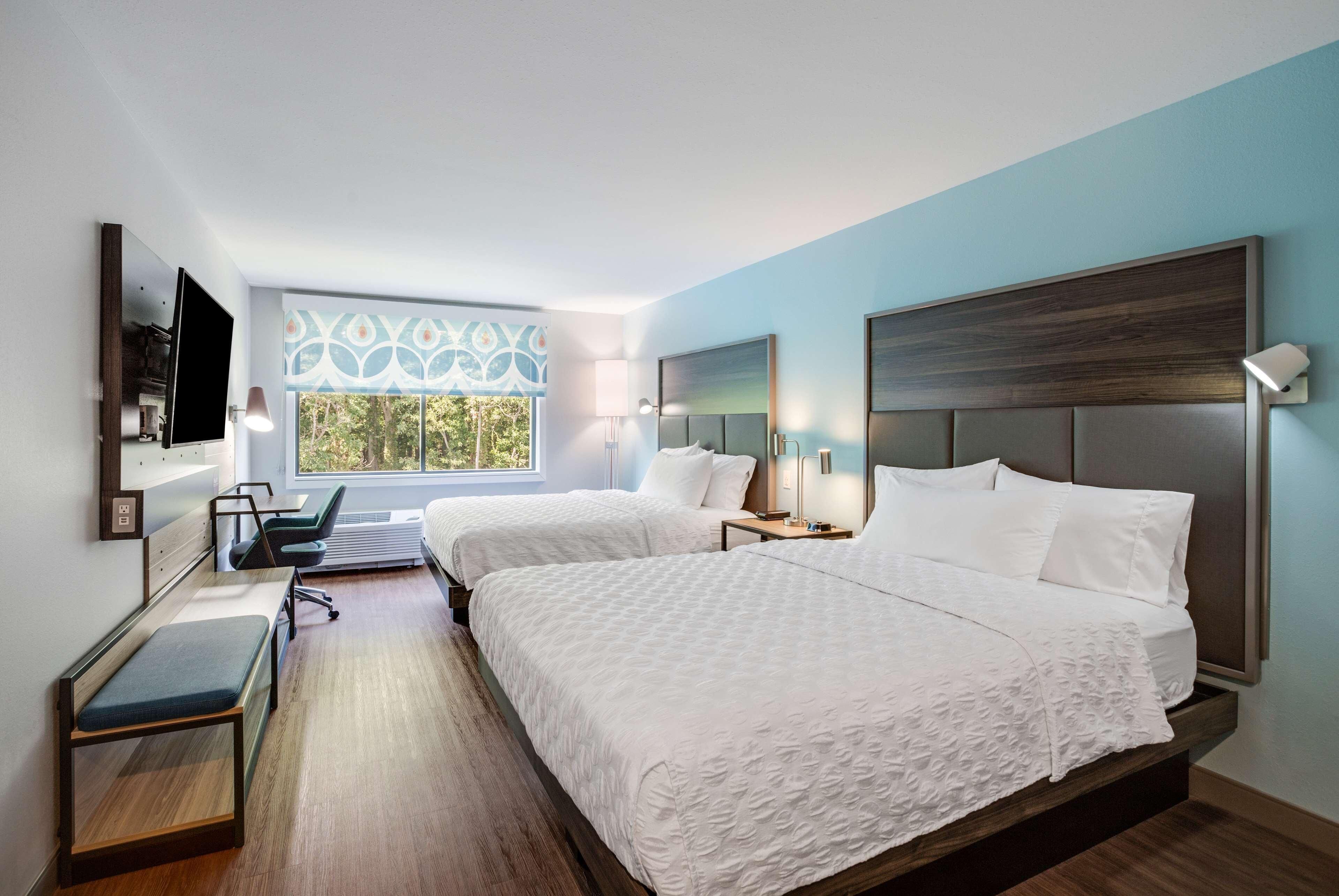 A hotel room with two beds, a desk, a chair, a TV, and a window with a view of trees.