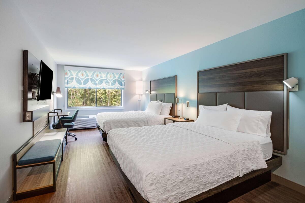Une chambre d’hôtel avec deux lits, un bureau, une chaise, une télévision et une fenêtre donnant sur des arbres.