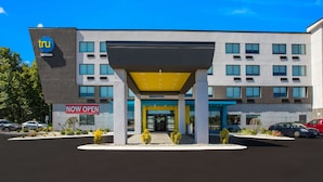 Een modern gebouw met een 'Nu Geopend'-bord, met een centrale ingang met een geel markies, meerdere ramen en een parkeerplaats met auto's.