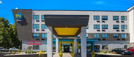 Ein modernes Gebäude mit einem 'Now Open'-Schild, das einen zentralen Eingang mit einem gelben Vordach, zahlreiche Fenster und einen Parkplatz mit Autos bietet.