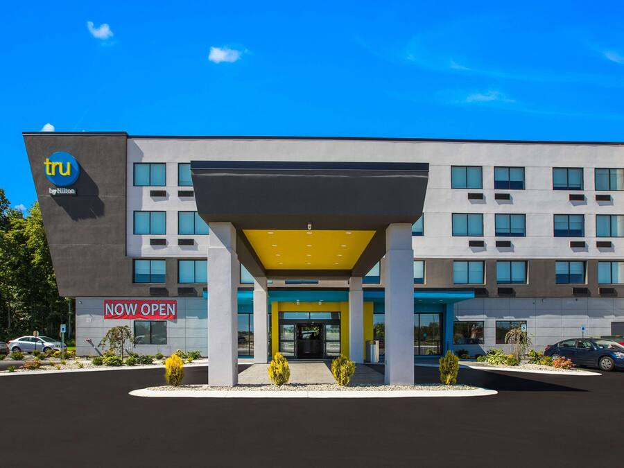 A modern building with a 'Now Open' sign, featuring a central entrance with a yellow canopy, multiple windows, and a parking lot with cars.