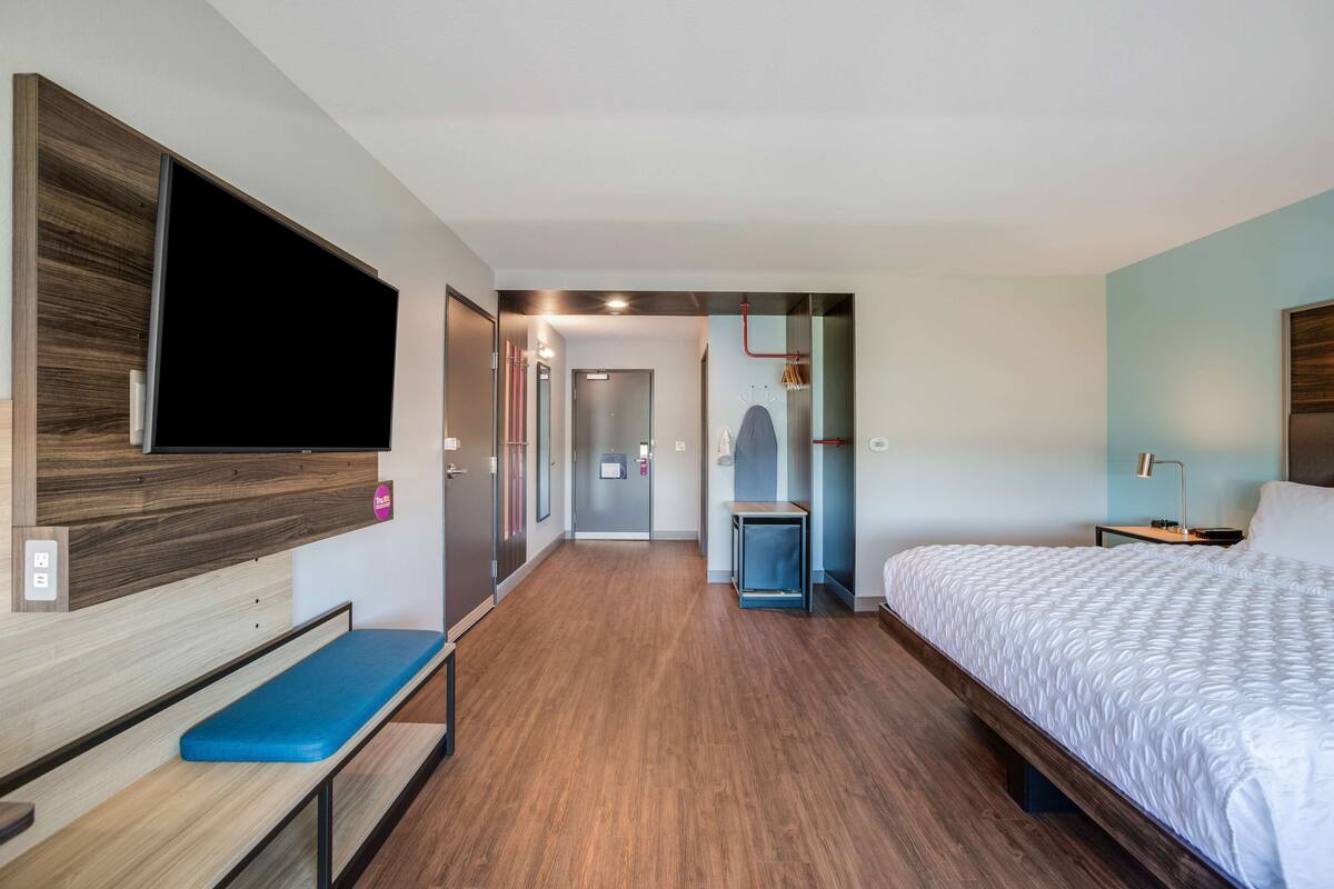 Une chambre d’hôtel moderne avec un banc en bois, un grand lit, un téléviseur fixé au mur et un couloir menant à la salle de bain.