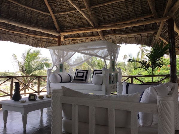 A white, thatched-roof outdoor seating area with a canopy, white cushions, and a small table with a vase and a bowl.