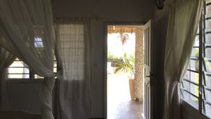 Ein Zimmer mit Bett, einem Fenster mit durchsichtigen Vorhängen, einer zur Außenseite führenden Tür und einer Steinsäule.