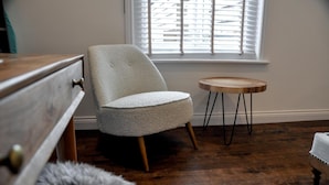 A room with a wooden floor, a white chair, a small round wooden table, and a window with blinds. - Upstairs at 33 (Worcester)