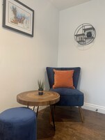 Una habitación de esquina con un sofá azul, una mesa redonda de madera con una planta, y un cuadro en la pared.