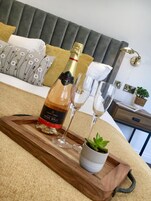 Uma cama com cabeceira estofada cinza, uma bandeja de madeira com uma garrafa de champanhe, duas taças de vinho e uma pequena planta em vaso.