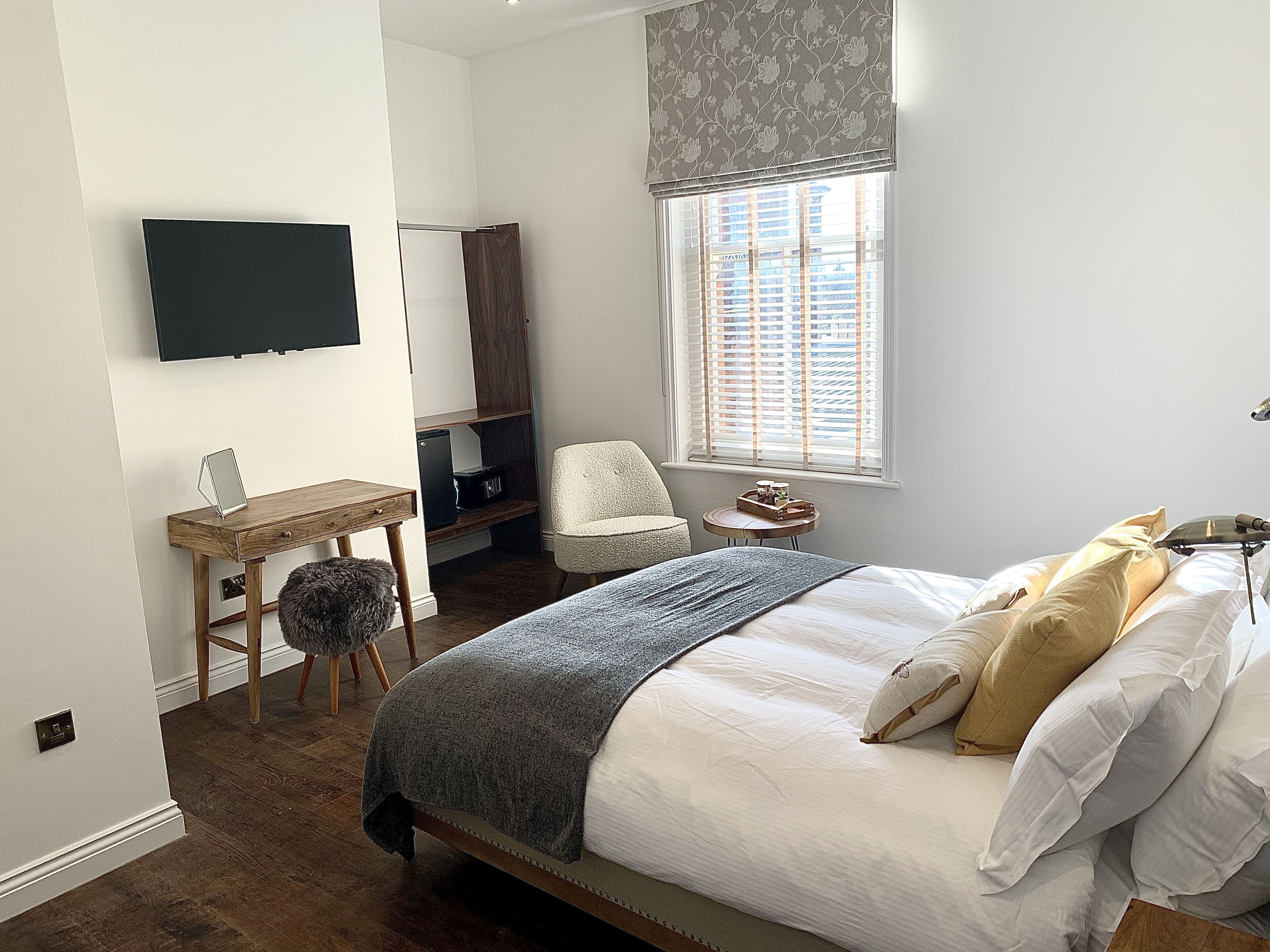 A bedroom with a bed, a desk, a chair, a TV, and a window with blinds.