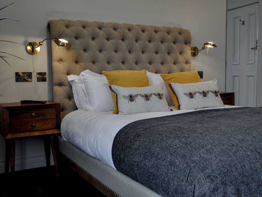 Een slaapkamer met een gestuukt hoofdeinde, een grijze deken en gele kussens. Er staan nachtlampjes en een houten nachtkastje met een lade.
