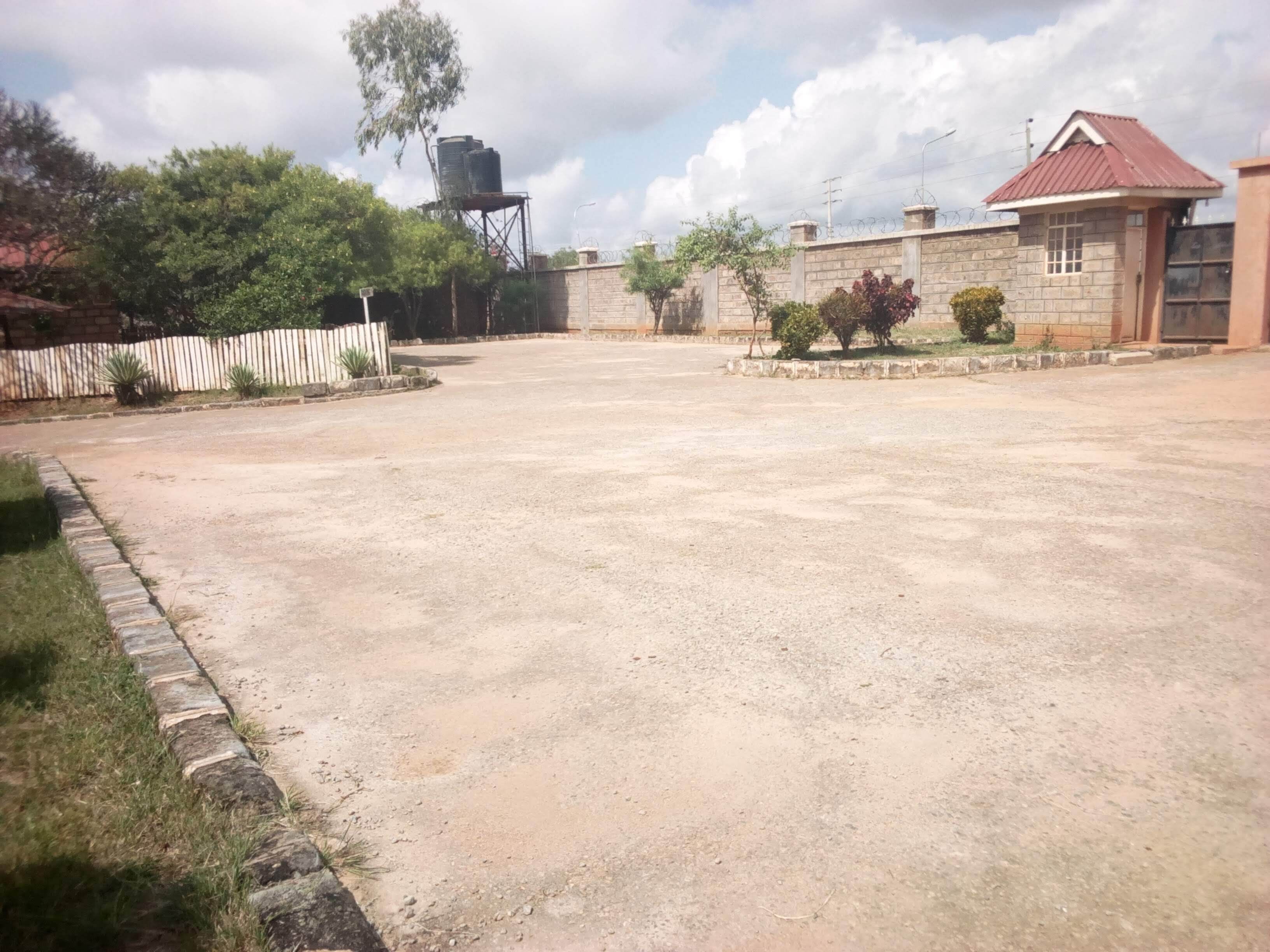 A spacious outdoor area with a paved ground, a fenced garden, and a building with a red roof.
