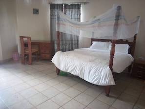 A bedroom with a four-poster bed, a wooden chair, a small table, and a curtain. - Mwingi Cottage Hotel (Mwingi)
