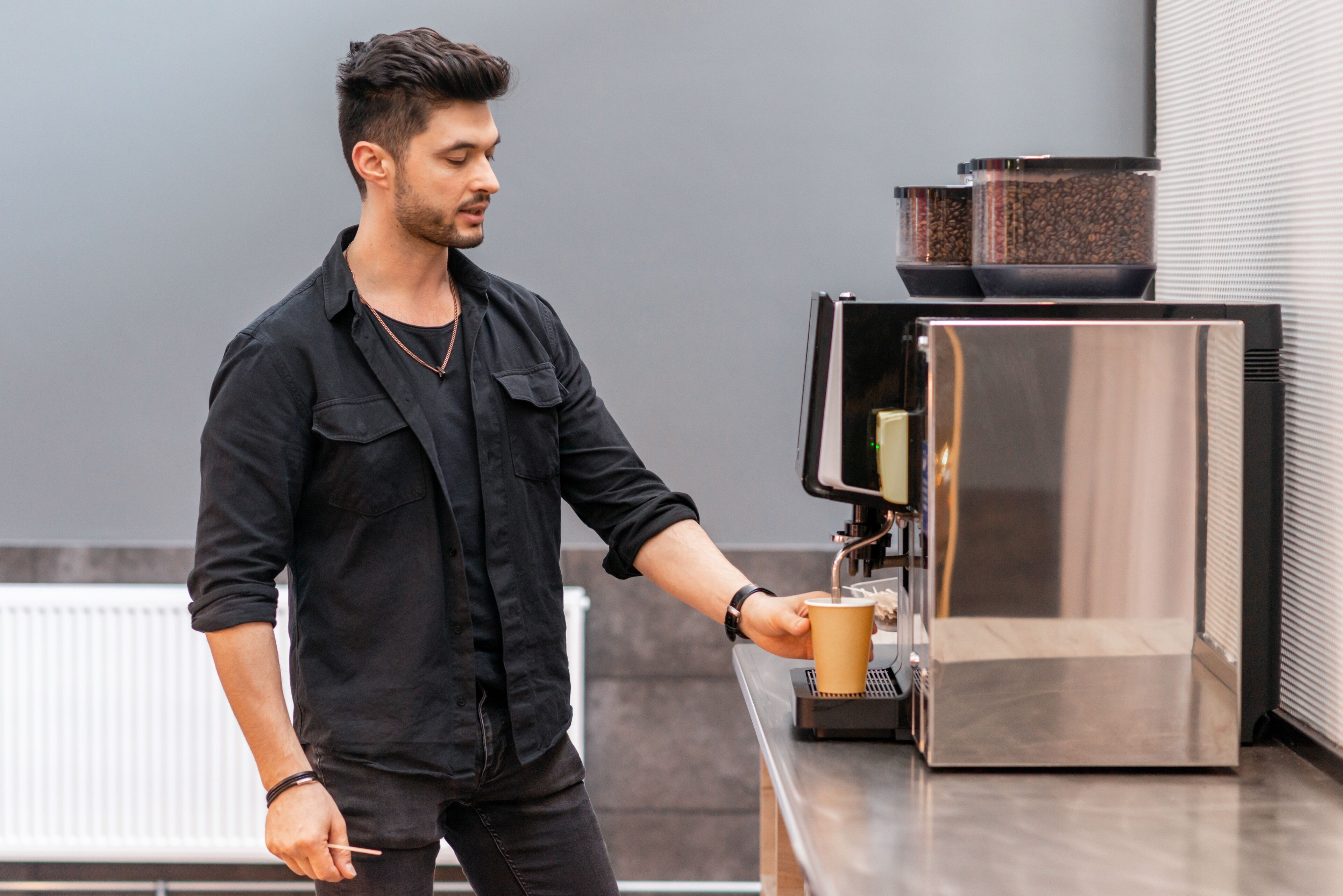 A person in a black shirt operating a coffee machine, with a cup in hand, in a modern kitchen setting.