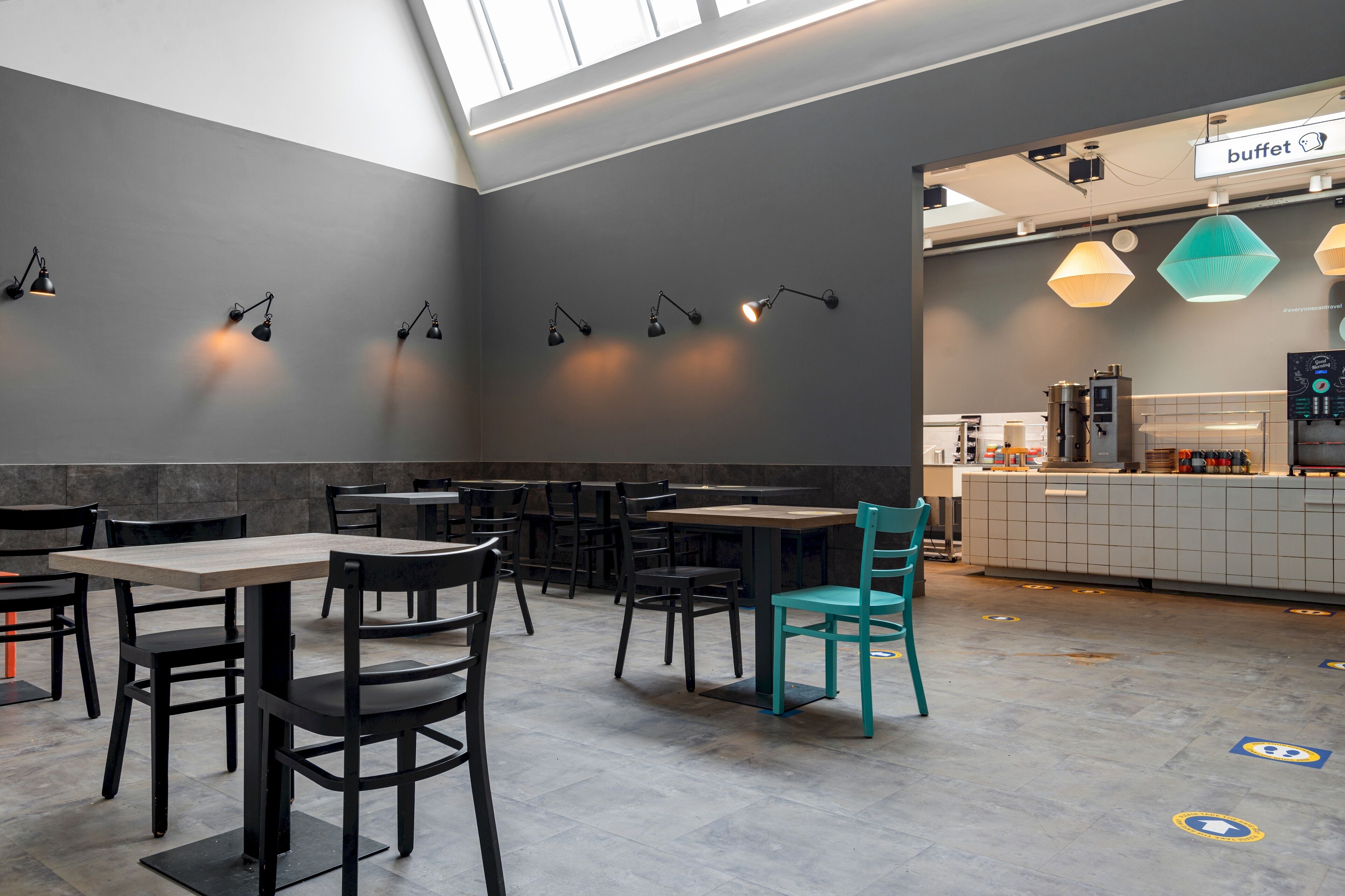 Et moderne restaurantinteriør med borde, stole, hængende lamper og et køkkenområde i baggrunden.