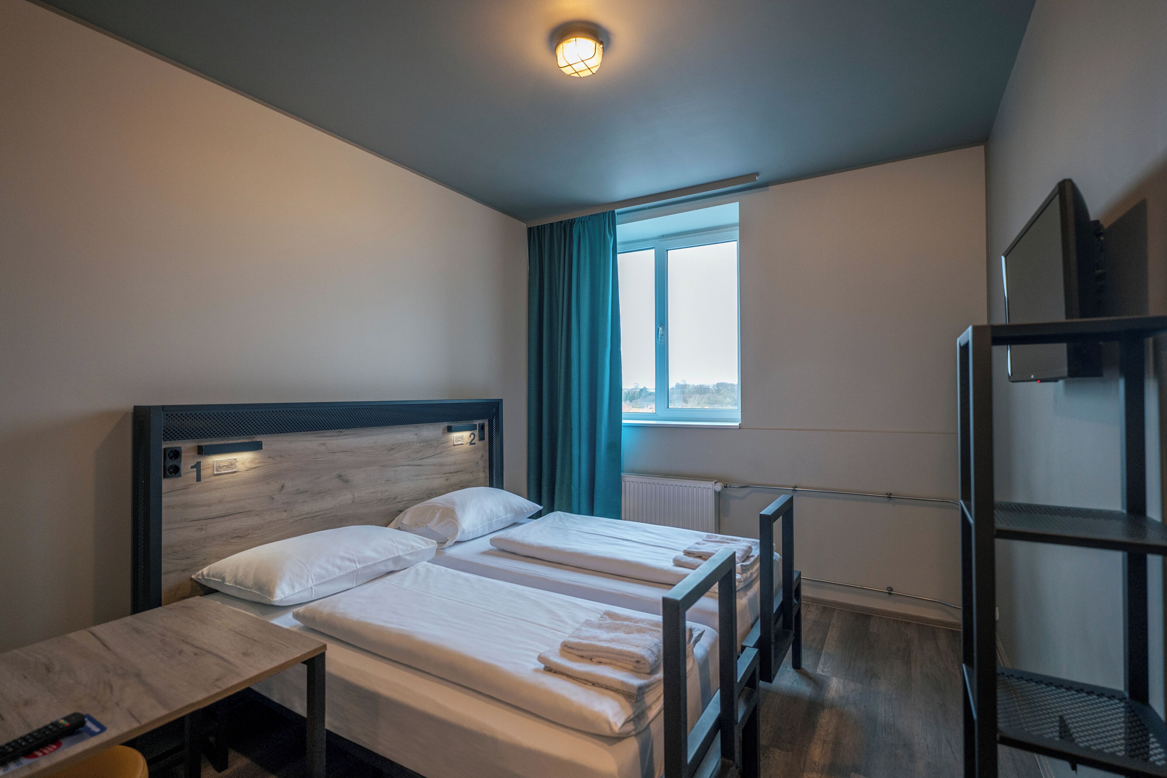 A hotel room with two beds, a desk, a window with curtains, and a bunk bed.