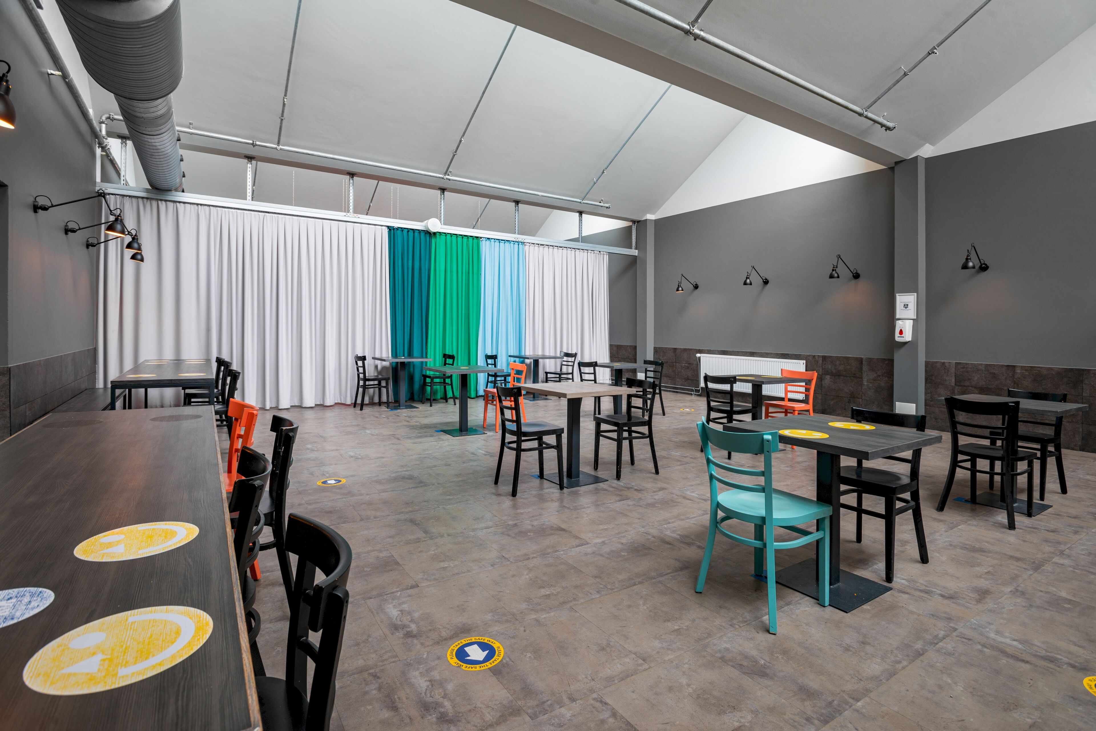 A modern dining area with tables and chairs, a high ceiling with exposed ductwork, and a large window with curtains.
