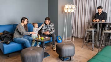 A living room with a blue sofa, a small round table, and a couple of ottomans. There is a baby on the sofa, a man sitting on the floor, and another man at a table.