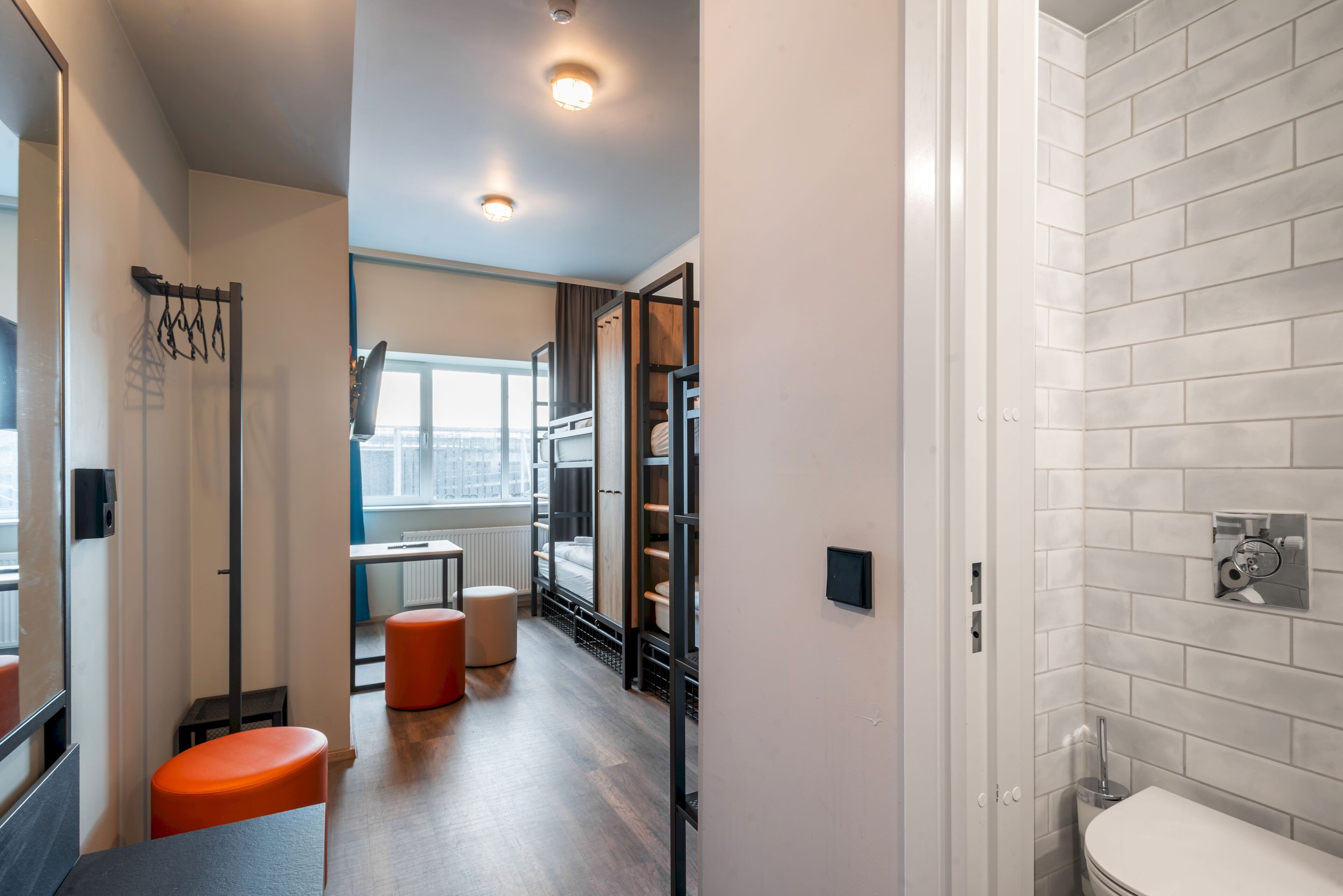 Habitación de hotel moderna con suelo de madera, ventana con vistas, baño con azulejos blancos y una zona de estar con taburetes naranja.