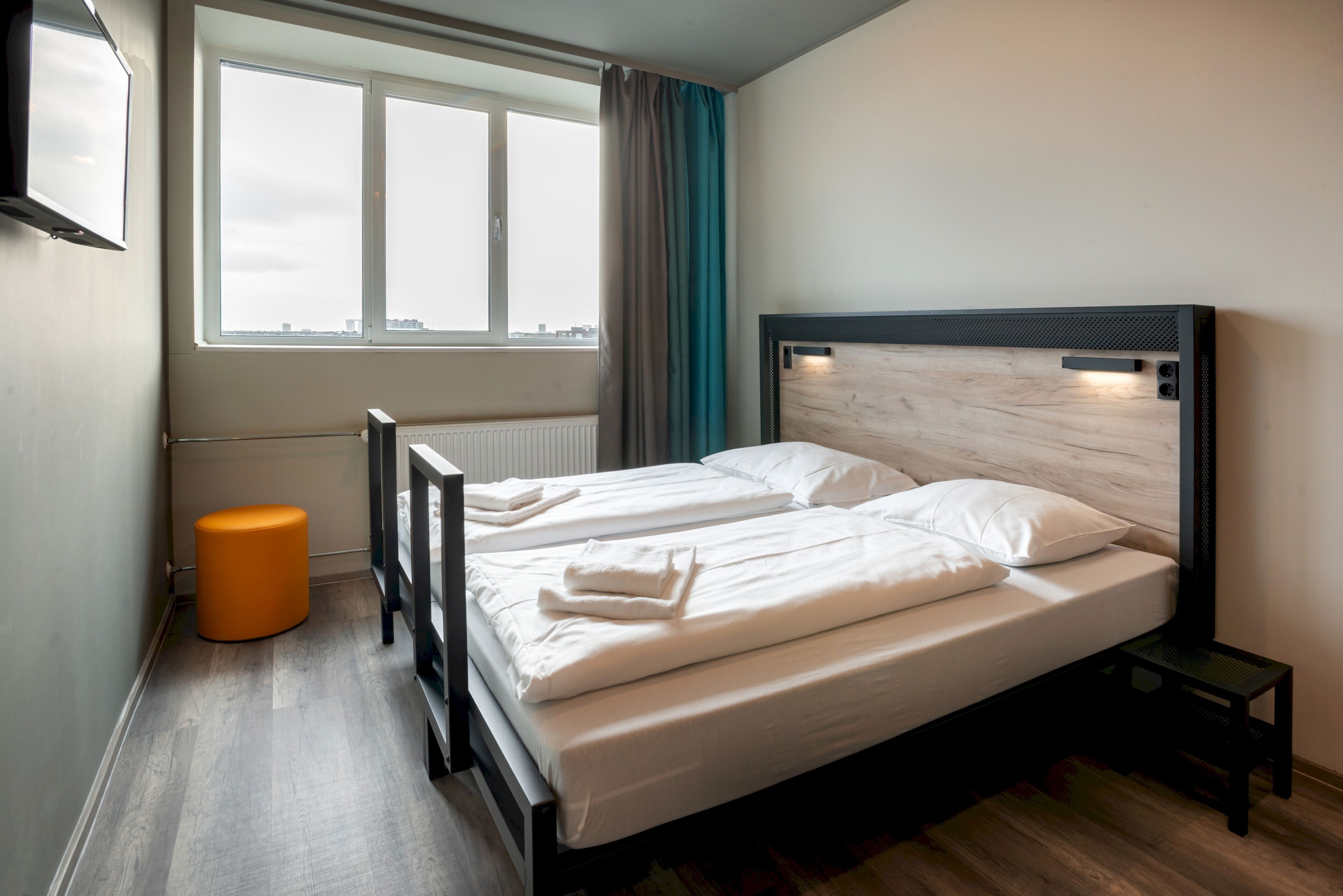 Quarto com duas camas, uma janela com cortinas, um radiador e um banquinho laranja pequeno.