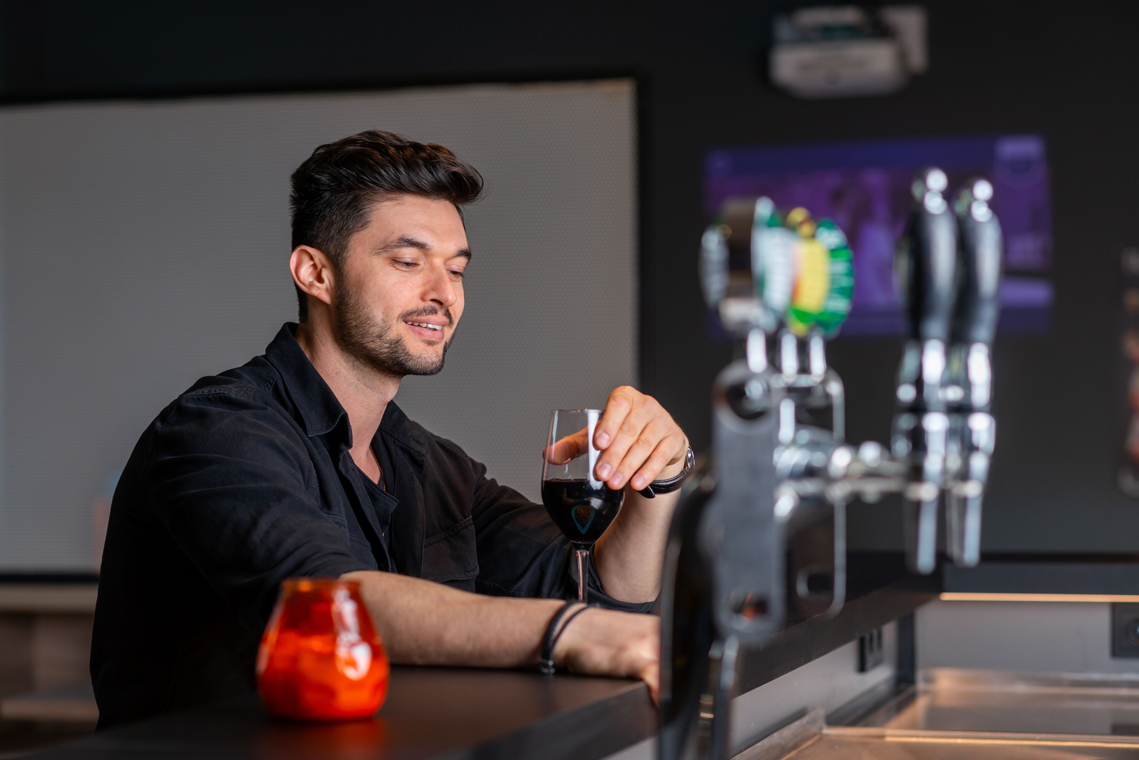 A man sitting at a bar with a glass of red wine, a beer tap, and a projector screen in the background.