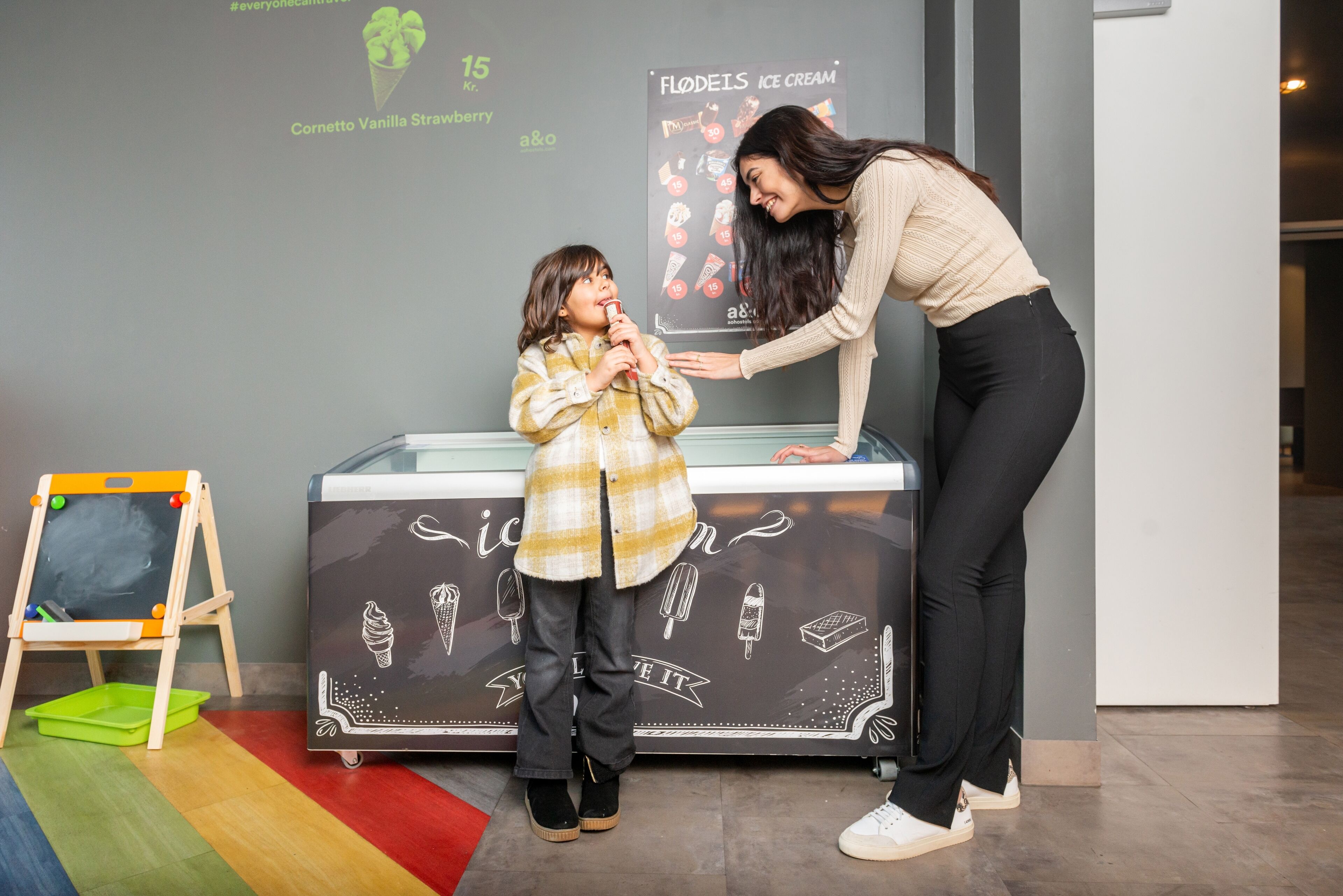 A child in a yellow plaid shirt and black pants standing in front of an ice cream display with a woman leaning over, possibly offering ice cream.
