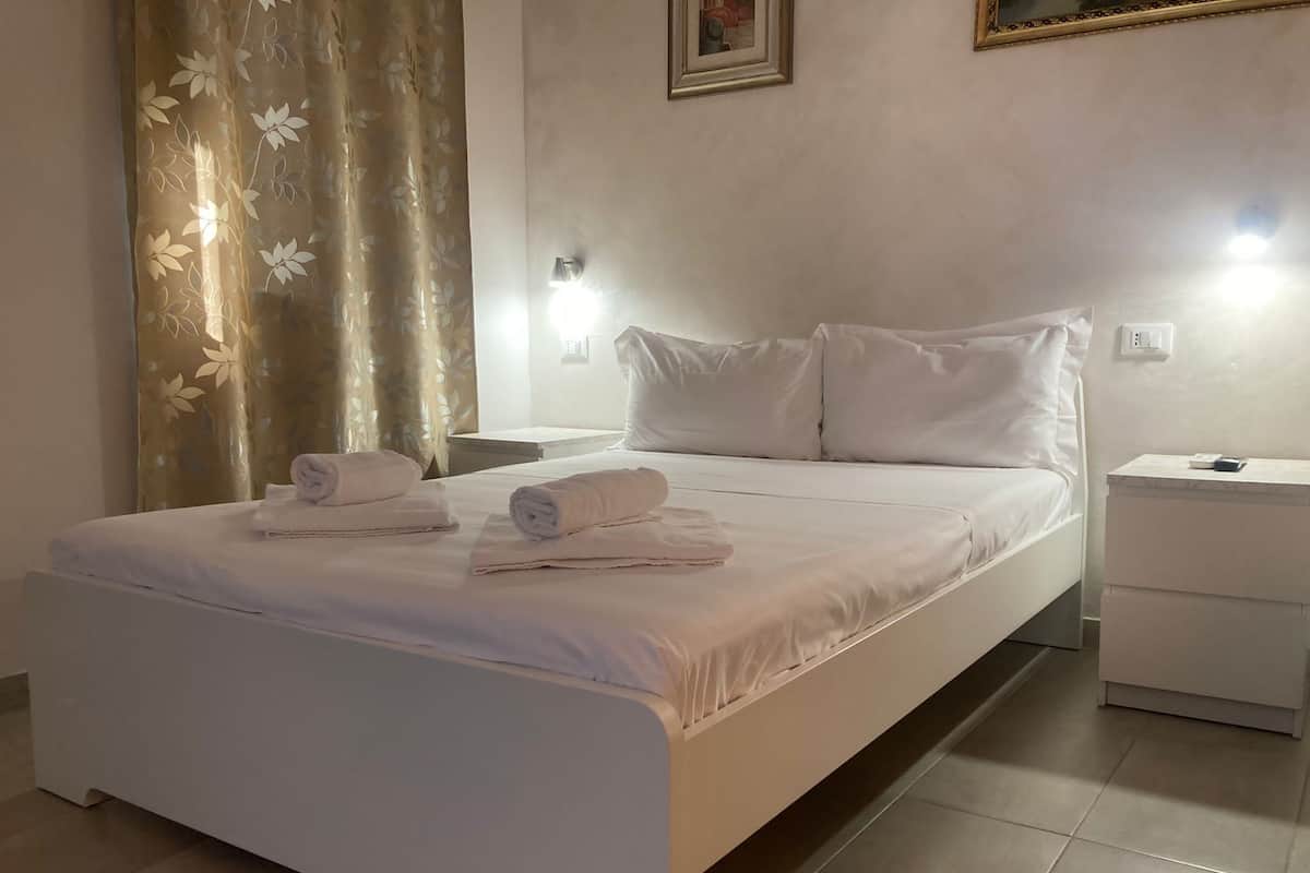 A neatly made bed with white linens and towels, a bedside table with a lamp, and a wall-mounted light fixture.