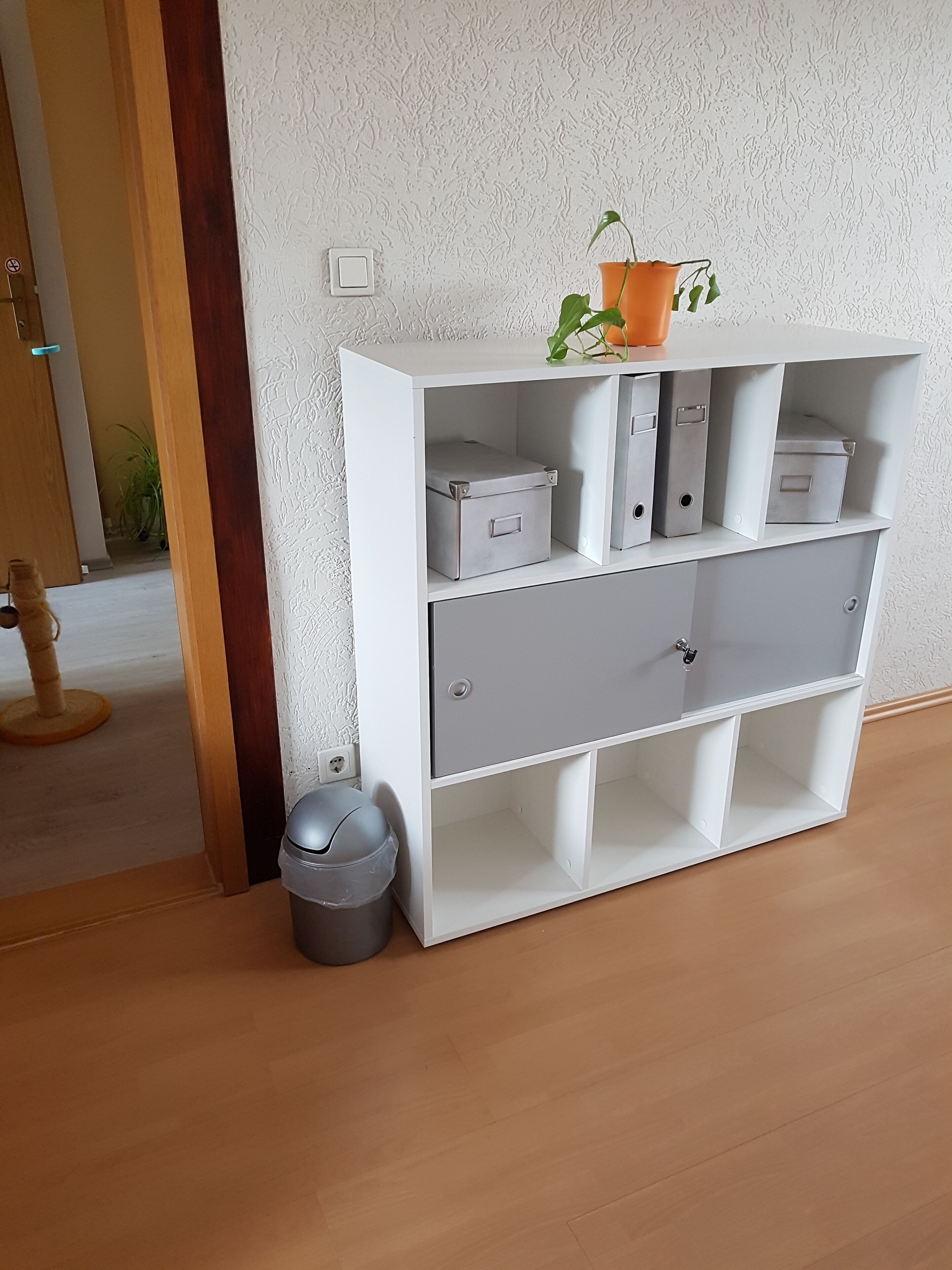 Een witte bergingsunit met verschillende vakken, een potplant en een prullenbak.