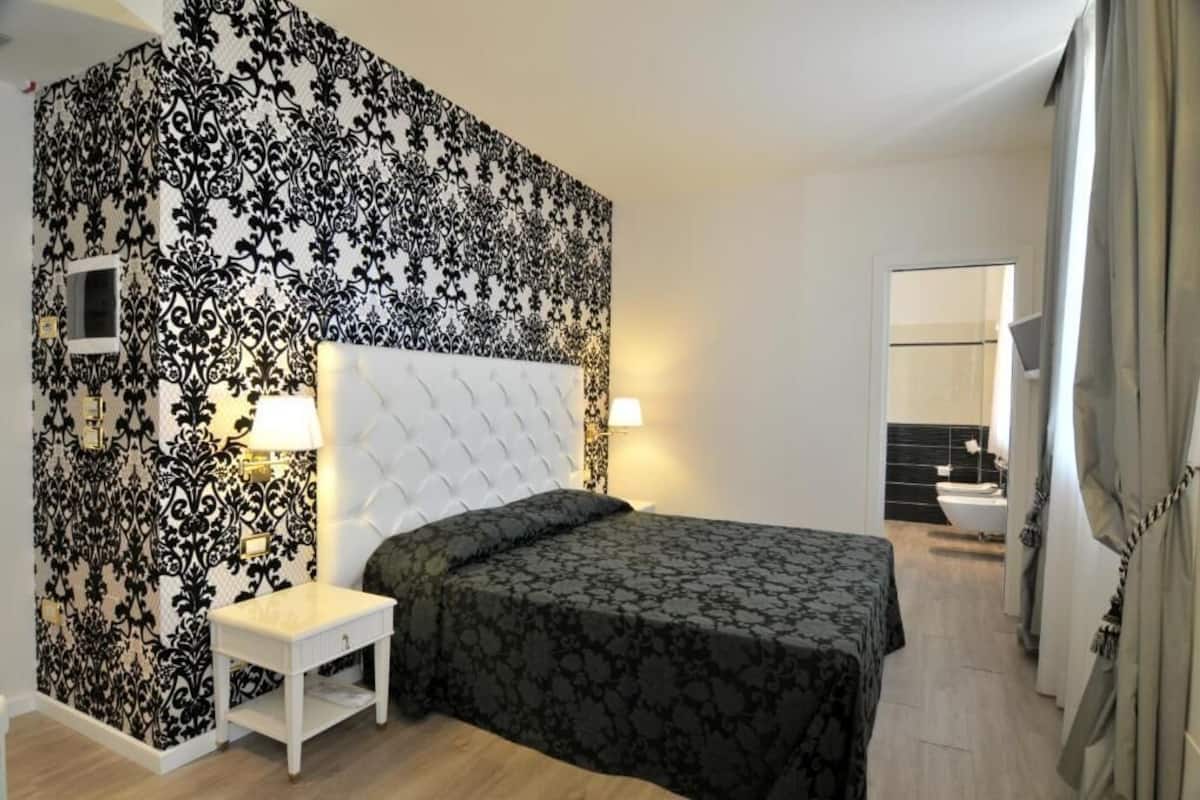 Une chambre à coucher avec une tête de lit capitonnée, une table de chevet, une couvre-lit à motifs et un mur orné d’un motif damassé noir et blanc.