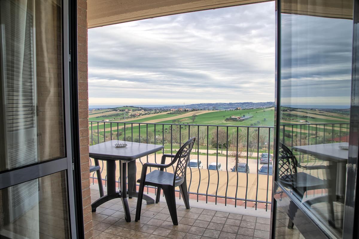 Un balcon avec une table et des chaises, d’où l’on peut admirer un parking et des champs situés au loin.