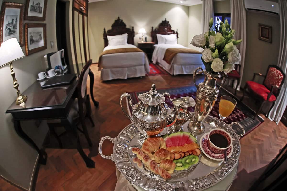 A hotel room with two beds, a dining table with breakfast items, and a desk with a lamp.