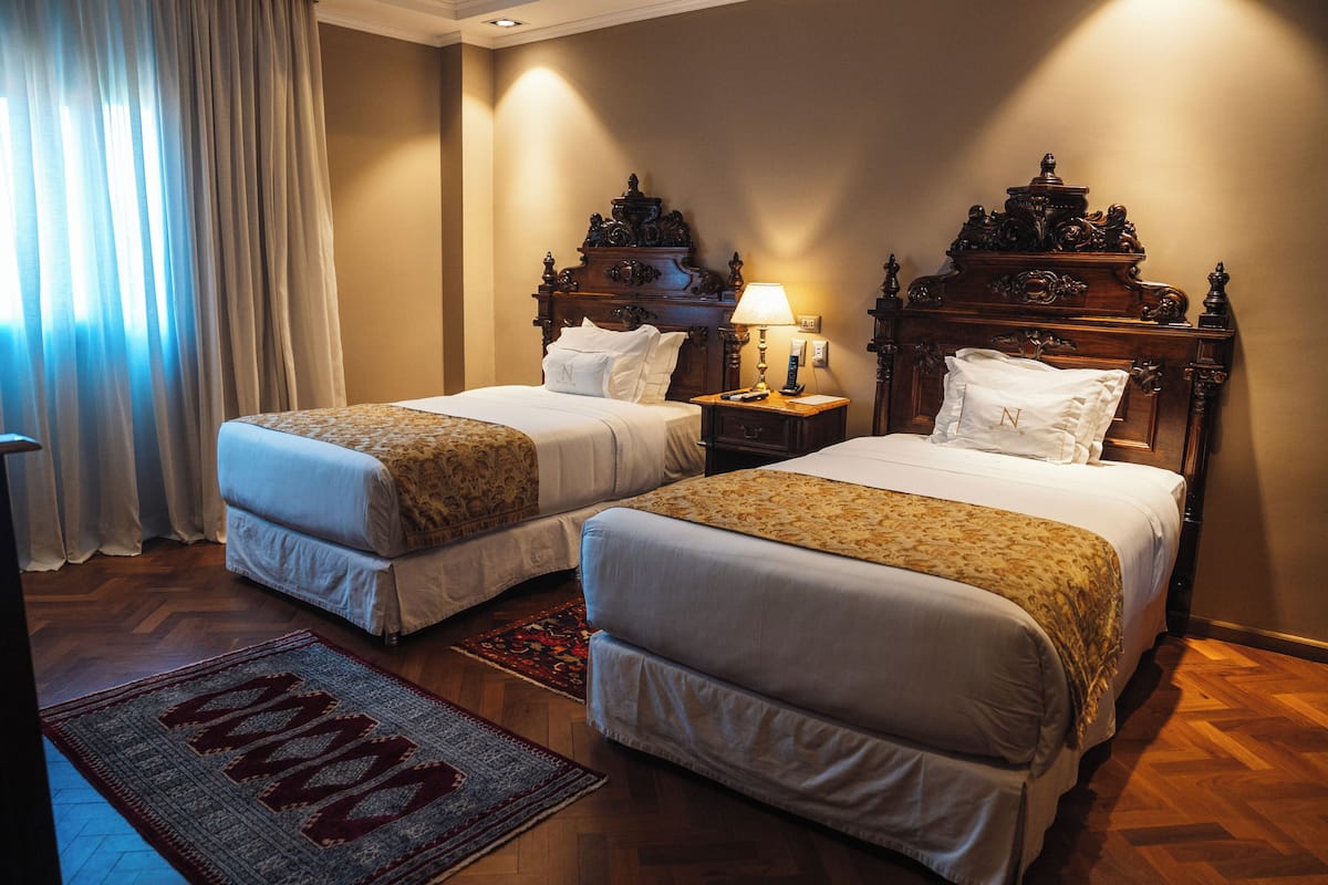 A hotel room with two beds, a nightstand with a lamp, a wooden headboard, and a patterned rug.