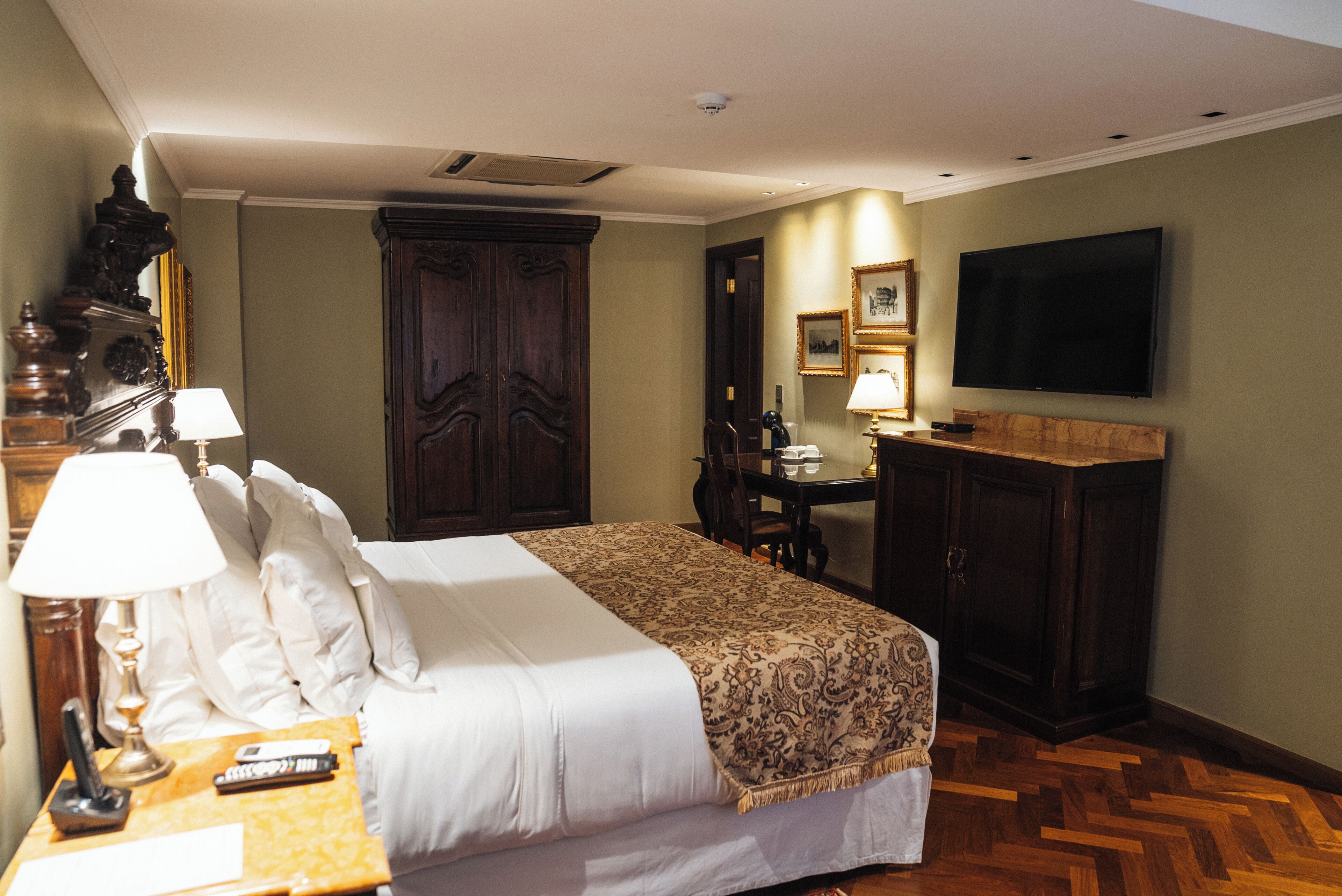 A hotel room with a bed, bedside table with lamps, a desk, a television, and a large wardrobe.