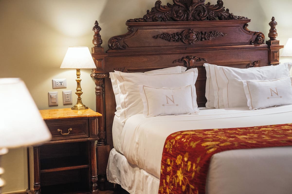 A neatly made bed with white linens and pillows, a wooden headboard, a nightstand with a lamp, and a red patterned throw blanket.
