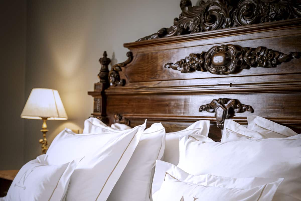 A neatly made bed with white linens and pillows, a wooden headboard with intricate carvings, and a bedside lamp.