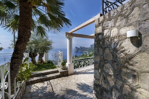 A stone pathway leading to a balcony with a white railing, a palm tree, and a view of the sea.