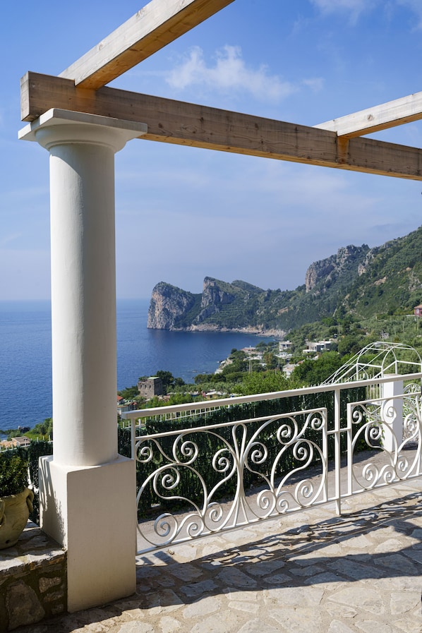A balcony with a white railing, a stone floor, and a view of the sea and mountains.