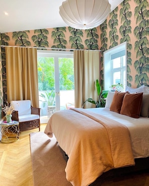 Un dormitorio con una cama grande, una silla, una mesita con libros y vistas al verdor a través de la ventana.