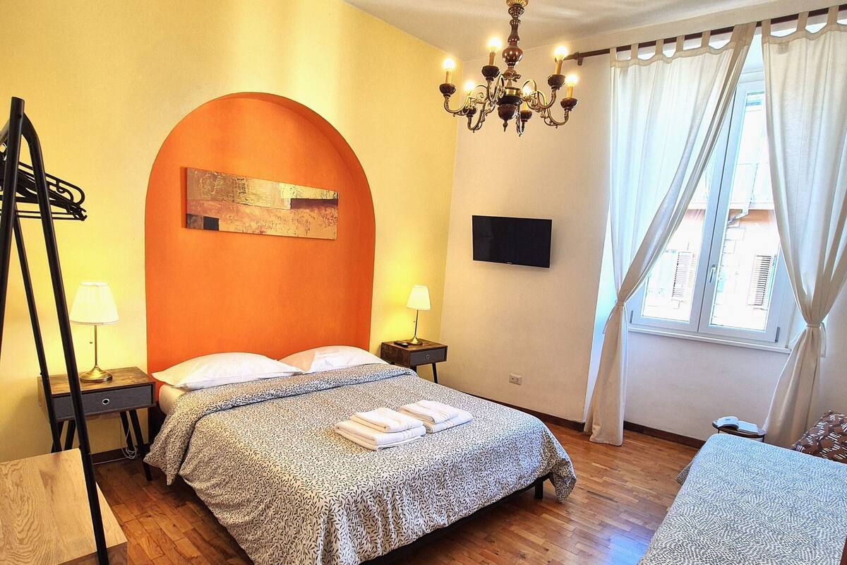 A hotel room with a bed, a chair, a TV, a chandelier, and a window with curtains.