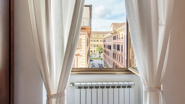Una ventana con cortinas blancas, vistas a edificios y un radiador en la parte inferior.