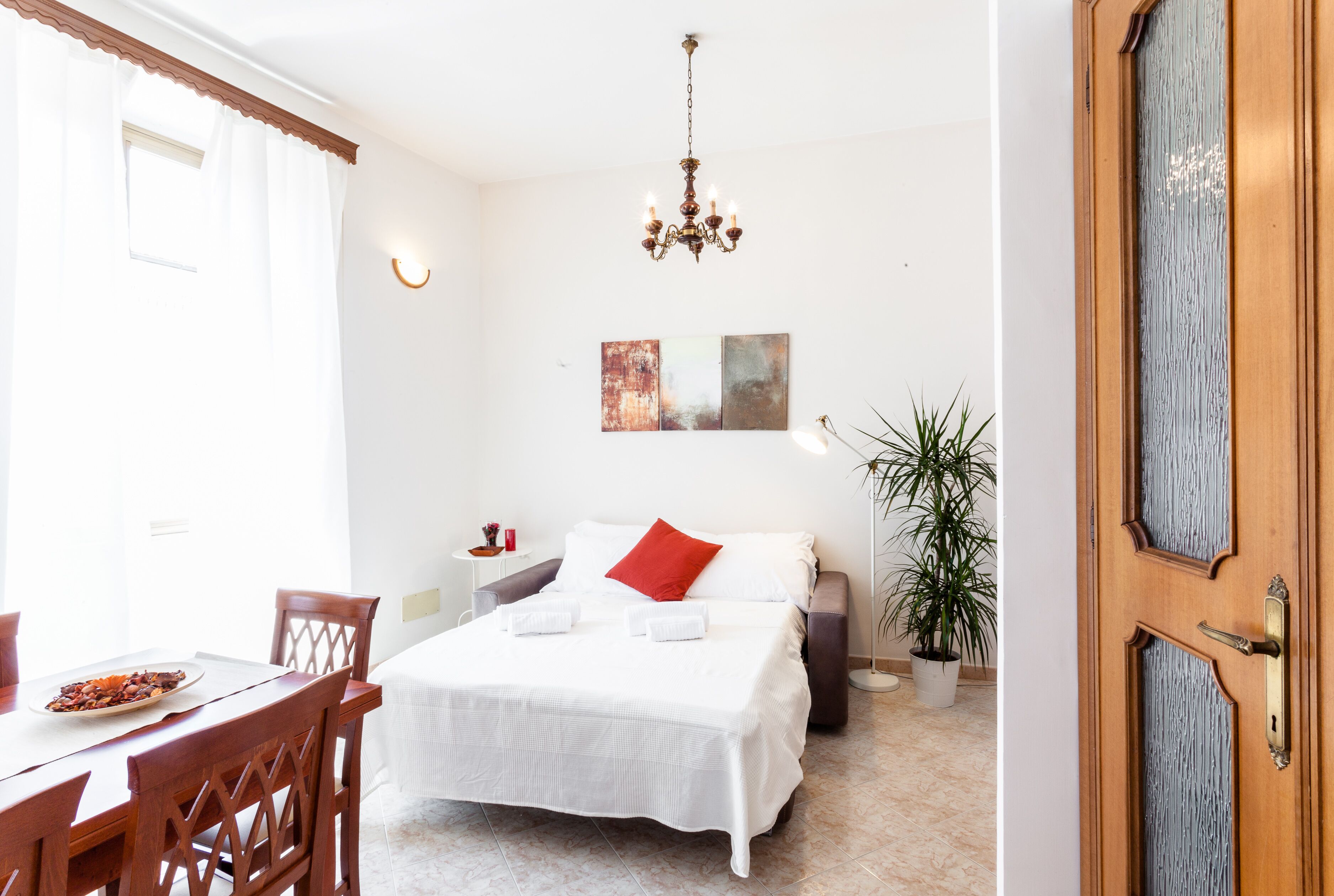 Habitación de hotel con cama, mesa de comedor, sillas, candelabro, obras de arte en la pared y una planta en maceta.