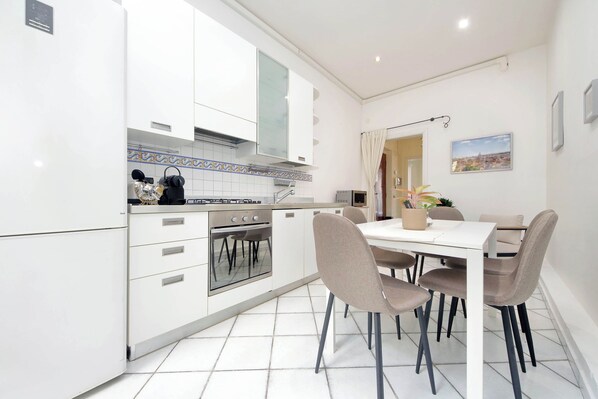 A modern kitchen with white cabinets, a stove, and an oven. There is a dining table with chairs and a vase with flowers. - Redseven Rome Collection (Rome)