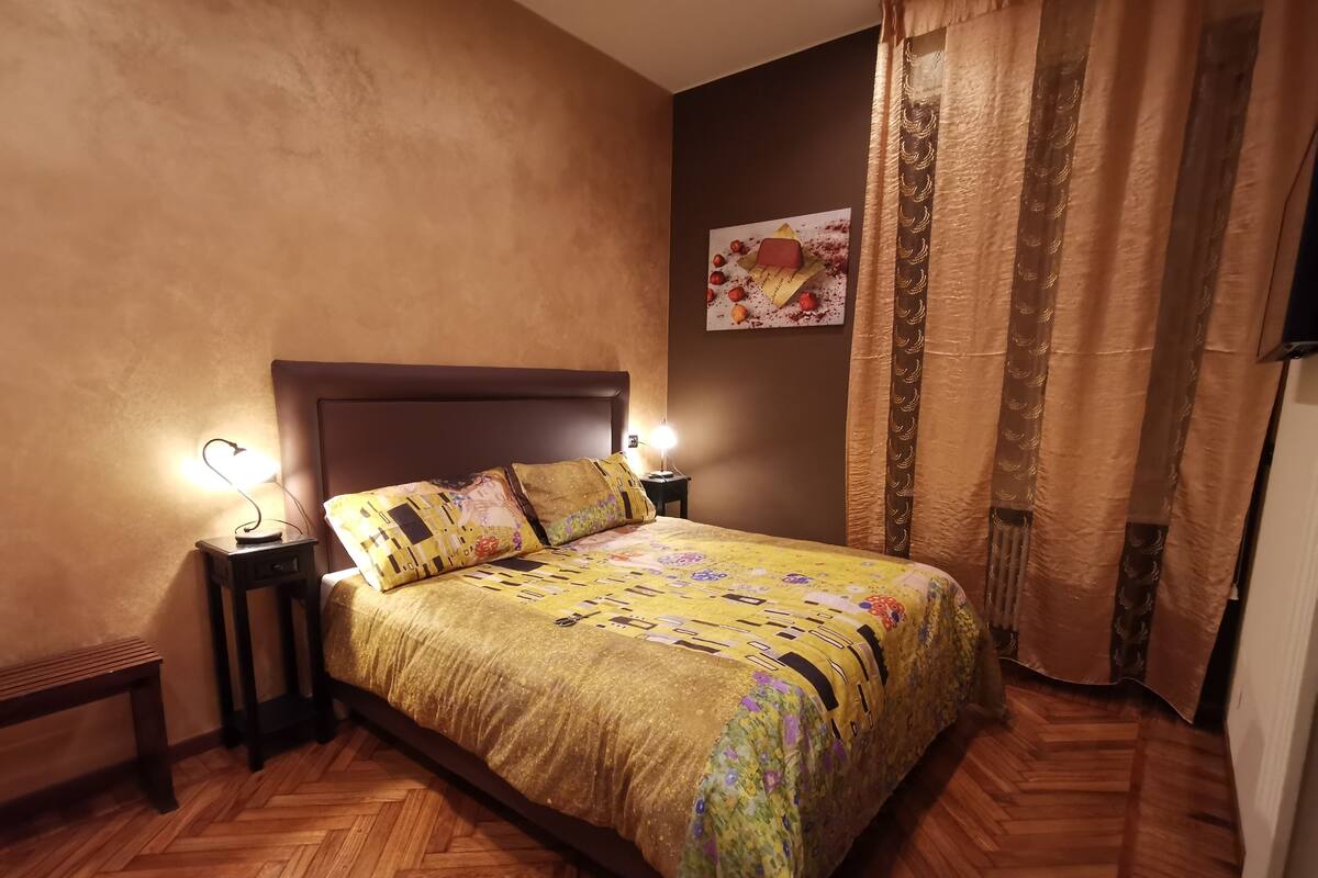 A bedroom with a large bed, two bedside lamps, a wooden bench, a patterned bedspread, and a wall-mounted artwork.