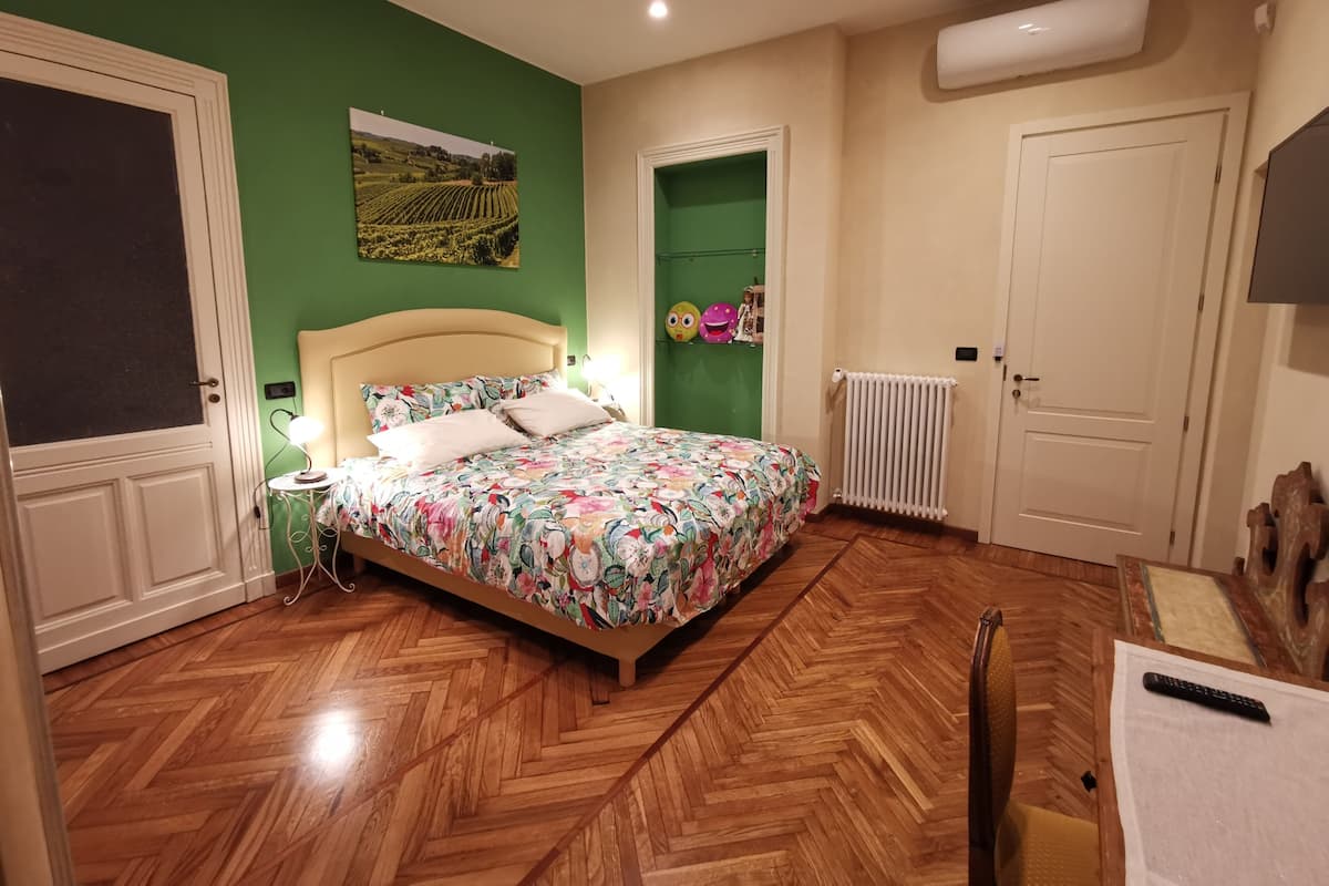 A bedroom with a wooden herringbone floor, a bed with a floral bedspread, a nightstand with a lamp, a radiator, and a door with a mirror.