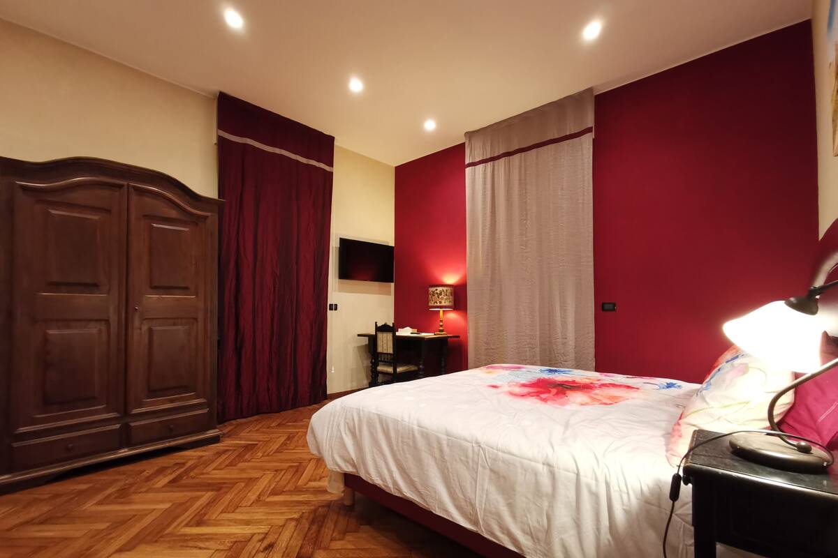 A bedroom with a wooden herringbone floor, a bed with a white bedspread, a desk with a lamp, and a wardrobe.
