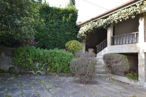 A stone building with a stone staircase leading to a balcony. There is a hedge and some flowering plants in front of the building.